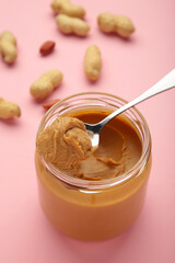 Jar and spoon of peanut butter on pink background.