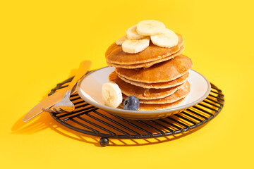Plate on grate with tasty pancakes, banana slices and cutlery on yellow background