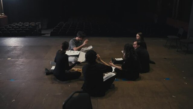 A confident young male director with script sheets in his hands tells young actors in black suits how to play their roles correctly during preparation and rehearsal for a performance on stage in a