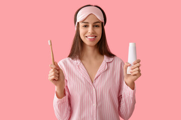 Young woman in pajamas and mask holding tooth brush and paste on pink background