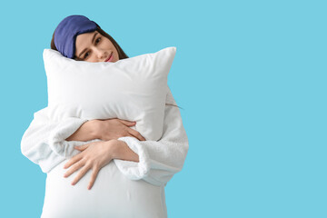 Young woman in pajamas and sleeping mask holding pillow on blue background