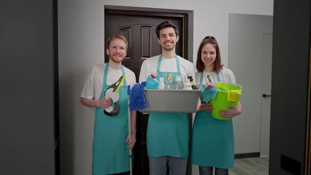 Portrait of a happy trio of cleaners in blue fronts with various tools and a cleaner for cleaning in their hands two men and a brunette girl in a modern apartment on a cleaning call