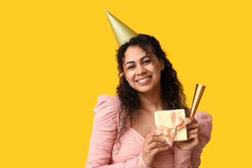 Beautiful young African-American woman in party hat with whistle and gift box on yellow background