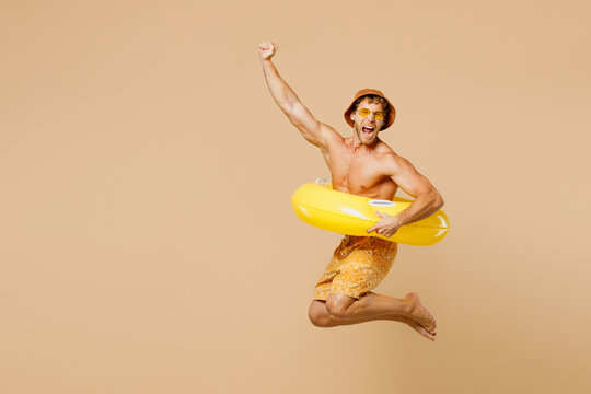 Full body young man wears yellow shorts swimsuit hat relax rest near hotel pool jump in inflatable ring do winner gesture isolated on plain beige background. Summer hot vacation sea sun tan concept.