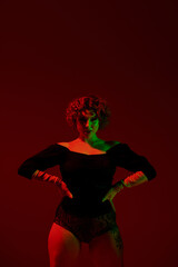 A young curvy redhead woman wearing a black dress, standing confidently in front of a striking red background.