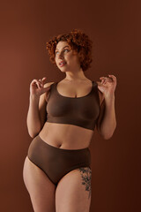 A young and curvy redhead woman in a brown bikini poses gracefully on a warm-toned brown background.