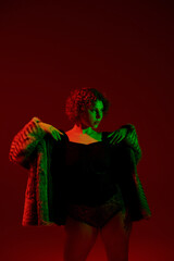 A young, curvy redhead woman strikes a pose in front of a vibrant red light.