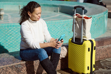Woman traveler sitting by a fountain using smartphone with luggage beside her