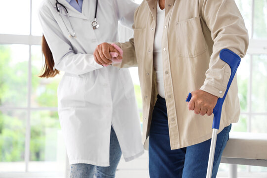 Female doctor helping mature man with crutch to walk in clinic