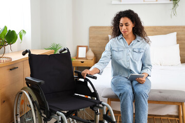 Young African-American woman with wheelchair and tablet computer sitting on bench at home