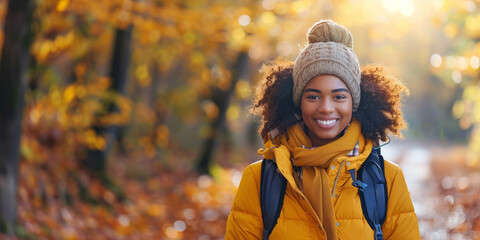 A young African American woman wearing a warm coat and hat smiles as she hikes through a forest in autumn