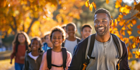 A happy African American male teacher leads a group of students on a field trip through the autumn leaves