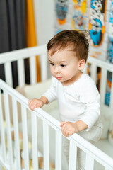 A baby is standing on a crib rail, looking at the camera. The crib is white and has a colorful design on the wall behind the baby