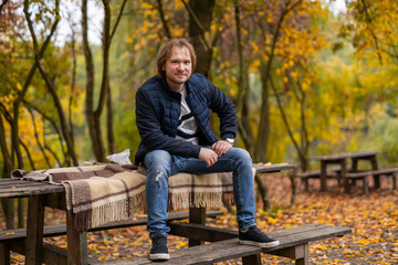 A man is sitting on a bench in a park. He is wearing a blue jacket and jeans