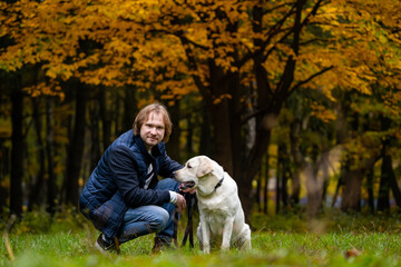 A man and his dog are standing in a grassy field with trees in the background. The man is wearing a blue jacket and jeans