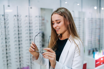 A woman is smiling and holding a pair of glasses in a store. The store is filled with many...