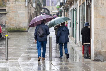 Two individuals are seen from behind walking down a wet cobblestone street, holding umbrellas to...