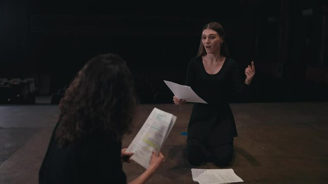 A confident girl with brown hair in a black theater costume plays out her role and tells how to read the words correctly during her rehearsal with a colleague on stage in an empty theater at a play