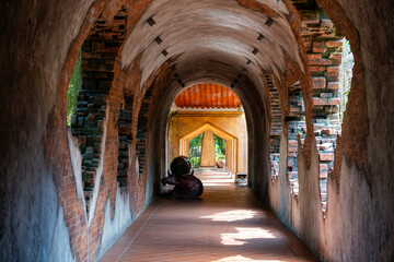 Fototapeta premium A rustic, arched corridor with exposed brick walls leads to a garden. Soft light filters through, casting shadows on the pathway. The scene is serene and peaceful, evoking a sense of history.