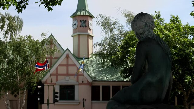 Silhouette of The Little Mermaid Fountain set against the backdrop of a traditional Danish building in Solvang, California. The flags of Denmark and Sweden flutter gracefully in the wind, adding a tou