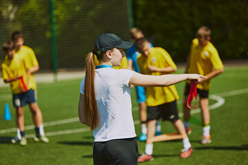 Fototapeta premium Group of young athletes, children training football outdoors with female coach on warm sunny day. Hobby and teamwork. Concept of sport, school, childhood, achievements, active lifestyle