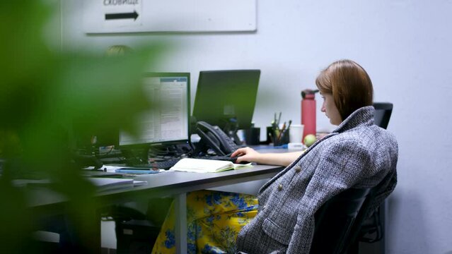 No heating girl in the office covered herself with a coat sitting working on computer. Working in the office is cold