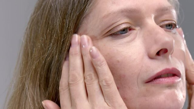 Old woman uses a moisturizer cream. Senior female applies anti-aging lotion with her hands and fingers, touches cheek, applying nourishing anti age face skin care. Wrinkles, pimples and acne on cheek