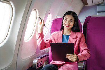 A young Asian female airplane passenger sits by the window during the flight, holding her boarding pass, with her carry-on luggage stored overhead, wearing her seatbelt, and enjoying the journey.