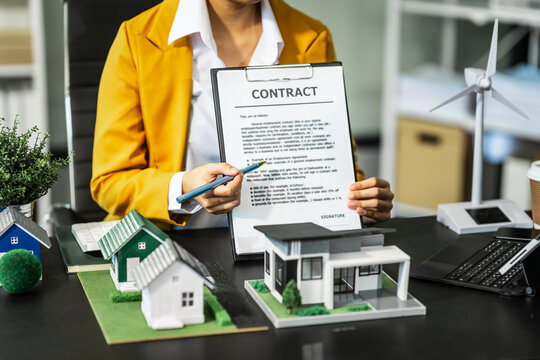 A businesswoman is reviewing contract papers at her desk in a sustainable house and condo, emphasizing clean energy with a nearby wind turbine powering the neighborhood.