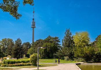 Obraz premium Vienna, Austria, August 17, 2022. The Danube Tower or Donauturm is the tallest building in Austria at 252 metres. It stands out among the trees of the park, three tourists walk along the avenue.