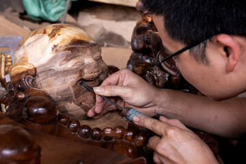 Photo of a young vietnamese man with glasses carving a wooden buddha head while working in the...