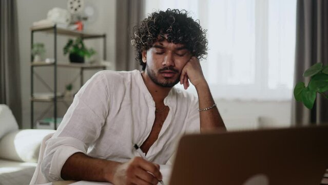 Tired exhausted stressed young indian man student preparing for exams and looking at scope of work with sad face at home workplace Overwork education concept	