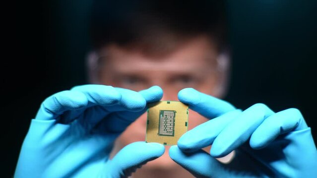 Selective focus shot of male engineer working in lab wearing protective goggles and gloves holding new processor in hands and examines it. Technician engineer male looking at detail for gadget.