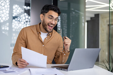 Happy Indian man sitting in office at desk, holding documents in hand, looking at laptop screen and...