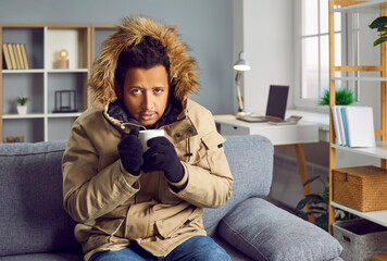 Young African man freezing at home in winter, sitting on the couch in a warm parka jacket and...