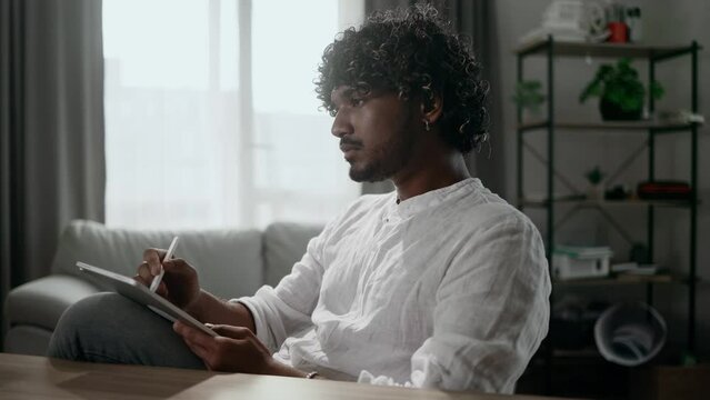 Handsome young indian man digital artist illustrator drawing sketch from nature using tablet in the light living room indoors Focused male painting by stylus at screen of digital pad at home workplace