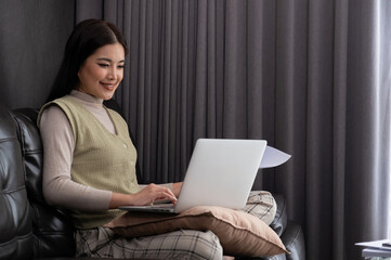 Smiling Asian woman using laptop on sofa, Concept of comfortable remote work