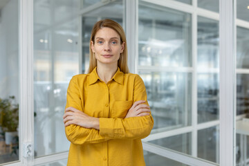Confident businesswoman wearing a yellow shirt stands with arms crossed in a modern office setting, looking directly at the camera