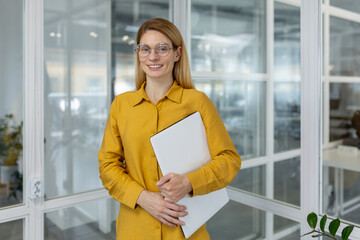 Confident businesswoman wearing yellow shirt and glasses, holding a laptop and smiling while...