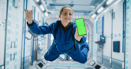 Female Astronaut Floating Inside a Space Station in Zero Gravity. Spacewoman Showing a Smartphone with Green Screen Mock Up Template. Woman Posing for Camera, Smiling in a Room in a Spacecraft