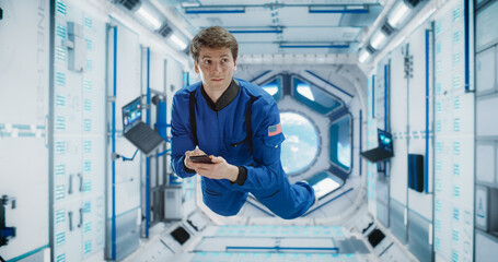 Cheerful Male Astronaut Floating in Zero Gravity Inside a Spaceship. Happy Caucasian Specialist Using Smartphone in Space, Checking News, and Communicating with Family and Friends Online.