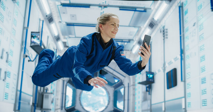 Cheerful Female Astronaut Floating in Zero Gravity Inside a Spaceship. Happy Caucasian Specialist Using Smartphone in Space, Checking News and Communicating with Family or Friends Online.