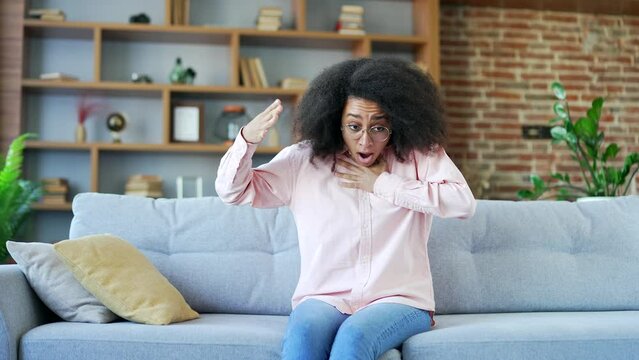 Young african american female having a panic attack sitting on sofa in living room at home. Black woman feels fear, restlessness and anxiety, holds her hands to her chest, breathes hard, is stressed