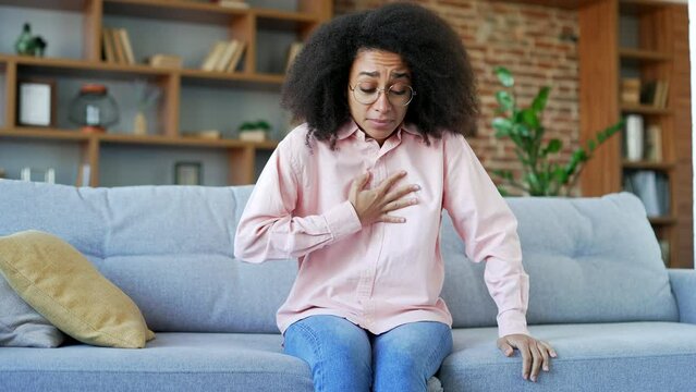 Sick young african american female suffering from chest tension, heart attack sitting on the sofa in living room at home. Black woman holding her chest with her hands, needs urgent medical attention