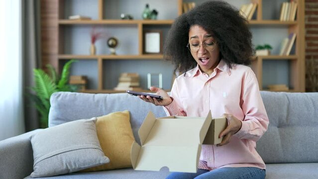 Disappointed african american female writes bad review on phone after receiving a parcel with wrong product at home. Black woman is unhappy with shopping in online store. Service issue or damaged item