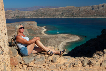 In the serene scene, a senior woman perches on a rock, gazing at a tranquil body of water. The azure sky above, she wears sunglasses, taking in the peaceful landscape of the sea
