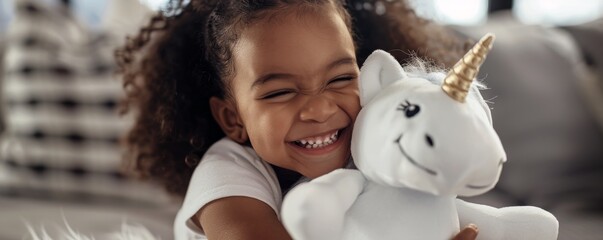 Little girl with afro hair style smiling and hugging unicorn plush toy at home on the sofa