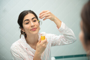 Fototapeta premium Young woman applying cosmetics on her face and looking at the mirror