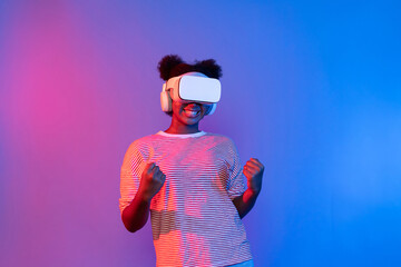 Young African woman with VR turning to metaverse playing game to be winner raising fist up neon...