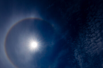 Obraz premium A captivating image of a sun halo against a deep blue sky, created by the refraction of sunlight through ice crystals in the atmosphere. The sun is bright, surrounded by a circular rainbow-like halo.